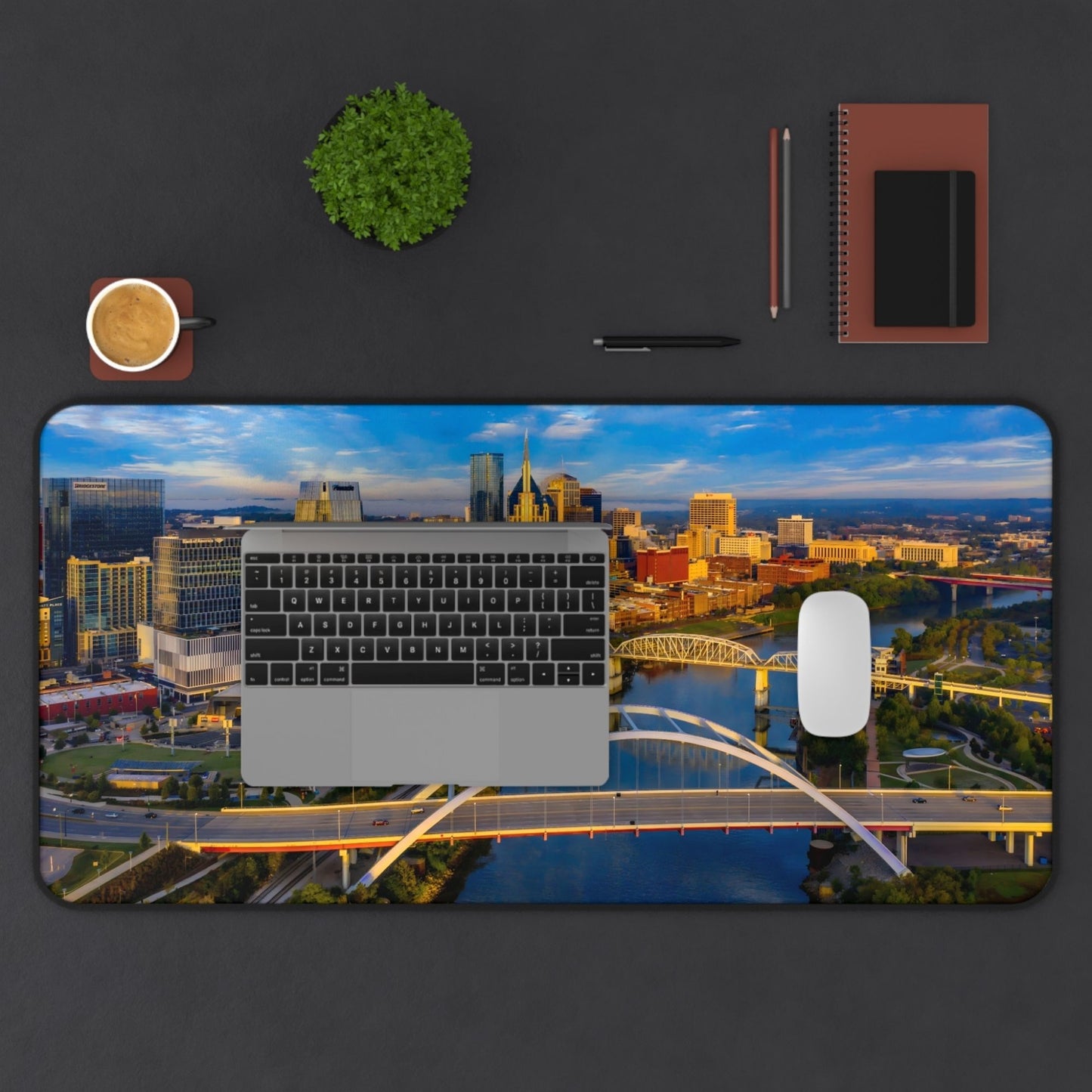 Desk pad 16 x 32 Desk setup with a cityscape mouse pad, laptop, cup, plant, pen, and notebook on a dark surface.